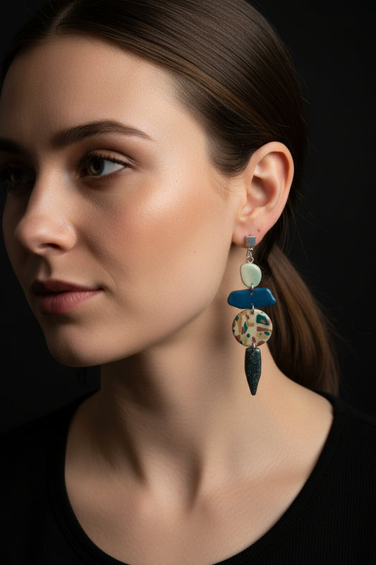 A pair of statement earrings on a white display card, photographed on a white marble-patterned surface. The earrings feature a blue translucent top bar with silver-toned cubes and two dangling silver-toned elements with a green, copper, and beige camouflage pattern.
